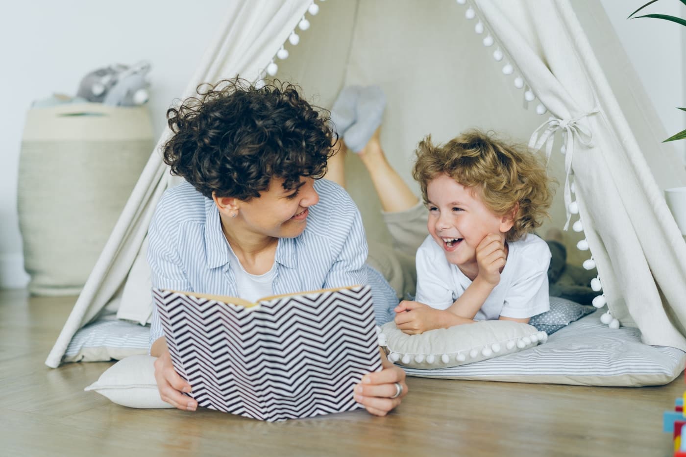 A parent lying on the floor reading with a laughing child inside a cozy play tent.